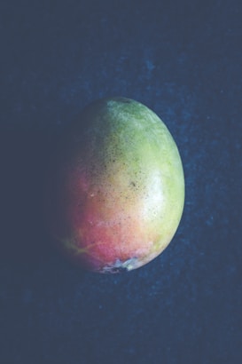 A single mango with a smooth texture and a mix of green, yellow, and orange-red hues rests against a dark, textured background. The lighting creates a soft, natural look with subtle shadows.