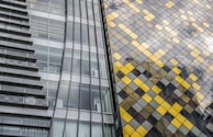 A modern architectural facade featuring a combination of glass panels and a decorative grid of yellow and grey tiles. The left side showcases a sleek glass exterior with visible horizontal beams, while the right side is adorned with a diamond pattern of shining yellow and muted grey squares. The structure reflects light, creating a dynamic and contemporary aesthetic.