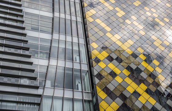 A modern architectural facade featuring a combination of glass panels and a decorative grid of yellow and grey tiles. The left side showcases a sleek glass exterior with visible horizontal beams, while the right side is adorned with a diamond pattern of shining yellow and muted grey squares. The structure reflects light, creating a dynamic and contemporary aesthetic.