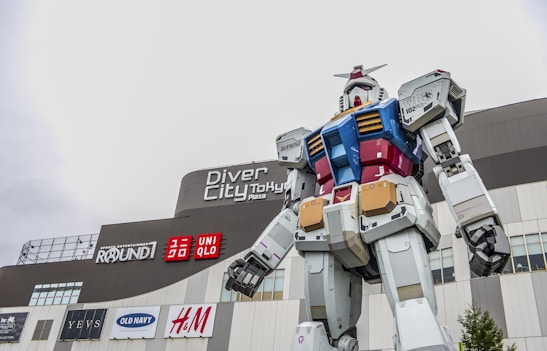A large robot statue stands prominently in an outdoor shopping complex. The robot is predominantly white with accents of blue, red, and gold. It has a distinct, mechanical design reminiscent of anime-style mechas. The backdrop features a modern building with various brand logos, including UNIQLO, H&M, and Old Navy.