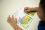 A child is reading a picture book with colorful illustrations of animals and a farmer in a natural setting. The child's hands are visible, holding the book open on a white surface.