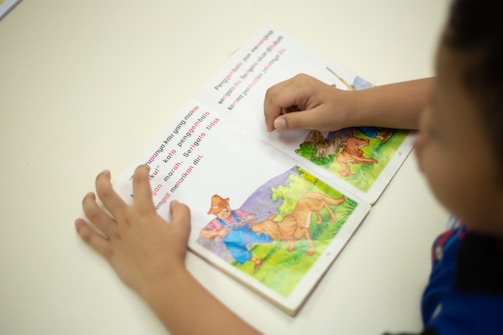 A child is reading a picture book with colorful illustrations of animals and a farmer in a natural setting. The child's hands are visible, holding the book open on a white surface.