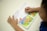 A child is reading a picture book with colorful illustrations of animals and a farmer in a natural setting. The child's hands are visible, holding the book open on a white surface.