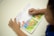 A child is reading a picture book with colorful illustrations of animals and a farmer in a natural setting. The child's hands are visible, holding the book open on a white surface.