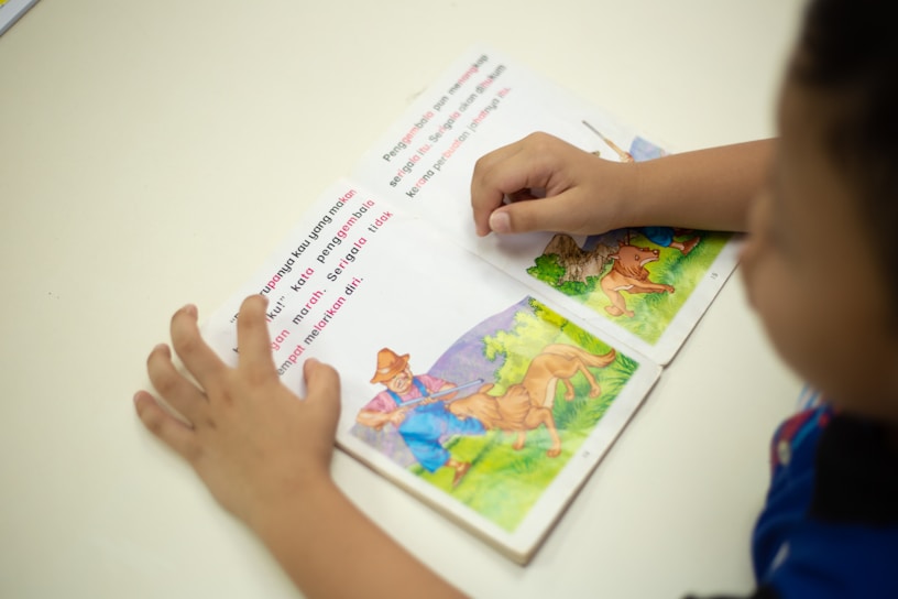 A child is reading a picture book with colorful illustrations of animals and a farmer in a natural setting. The child's hands are visible, holding the book open on a white surface.