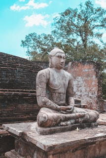 An ancient stone statue of a serene figure in a meditative pose is set against the backdrop of a weathered brick wall and lush green trees under a partly cloudy sky.