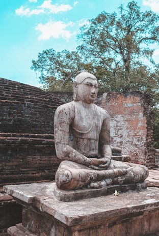 An ancient stone statue of a serene figure in a meditative pose is set against the backdrop of a weathered brick wall and lush green trees under a partly cloudy sky.