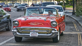 A vintage red Chevrolet convertible is parked on the street, likely from the late 1950s. The car has a Massachusetts license plate displaying the number 45674. Other modern vehicles and bicycles are visible in the background on a sunny day. Trees line the road, creating a mix of urban and natural settings.
