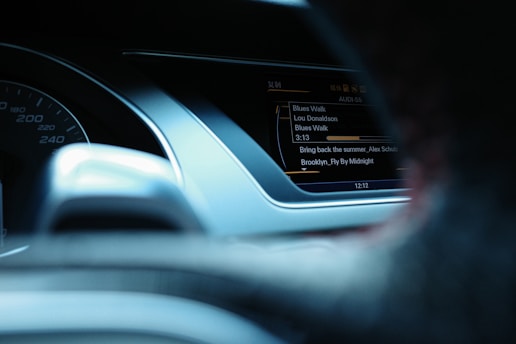 The interior of a car dashboard is visible, featuring a speedometer on the left side and a digital display on the right showing the music track information, including song titles and artists.