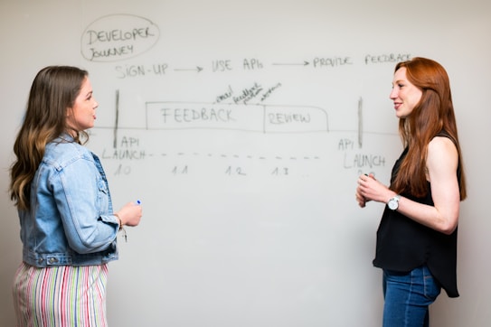 Two people are standing in front of a whiteboard, discussing a flowchart labeled 'Developer Journey'. The flowchart includes steps like 'Sign-Up', 'Use APIs', and 'Provide Feedback'. The individuals appear to be engaged in a professional discussion, with both focused on the content on the board. One person is wearing a denim jacket and holding a marker, while the other is in a black top.