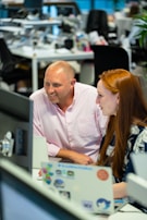 Two people are working together in an office environment. They are focused on a computer screen, smiling and engaged in a discussion. The workspace is modern, with desks, computers, and various office supplies in the background.