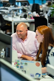 Two people are working together in an office environment. They are focused on a computer screen, smiling and engaged in a discussion. The workspace is modern, with desks, computers, and various office supplies in the background.
