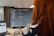 A person is sitting at a desk, focusing on a computer screen displaying code. The individual appears to be programming or debugging. The setup includes a large monitor and a laptop, surrounded by muted office lighting.