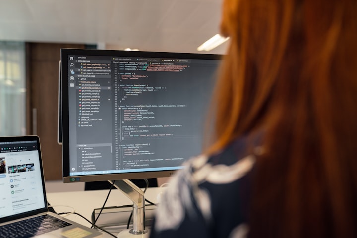 A woman sitting at a desk with a coding IDE open on her monitor.