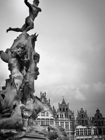 A dramatic black and white image featuring a detailed sculpture of a muscular figure poised in a dynamic stance at the top of a complex statue. The statue is adorned with other figures and mythical creatures in various poses. Behind the sculpture are historic European-style buildings with ornate facades and steep gabled roofs under a cloudy sky.
