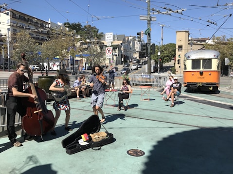 A street band performs music outdoors, consisting of three musicians playing an upright bass, a guitar, and a violin. People are sitting on outdoor chairs enjoying the performance under a clear blue sky. An orange and white streetcar is visible in the background, along with urban buildings and trees.