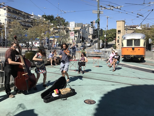 A street band performs music outdoors, consisting of three musicians playing an upright bass, a guitar, and a violin. People are sitting on outdoor chairs enjoying the performance under a clear blue sky. An orange and white streetcar is visible in the background, along with urban buildings and trees.