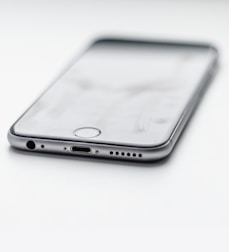 A close-up view of a smartphone lying on a flat surface, showcasing its charging port and headphone jack. The phone appears sleek with a metallic edge and is oriented at an angle.