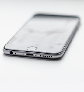A close-up view of a smartphone lying on a flat surface, showcasing its charging port and headphone jack. The phone appears sleek with a metallic edge and is oriented at an angle.