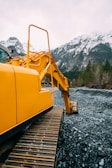 A large yellow excavator is positioned on rocky terrain with its arm extended. In the background, there are snow-covered mountains and a mix of evergreen trees. The sky is overcast, suggesting a cold and remote environment.