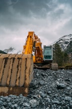 A large yellow excavator stands prominently on rough, rocky terrain with a majestic backdrop of snow-capped mountains and cloudy skies.