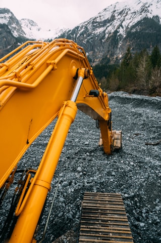 A large yellow excavator is positioned on a rocky terrain with snow-capped mountains and pine trees in the background. The metal arm of the excavator is extended prominently, showcasing the machinery's hydraulic components against a natural landscape.