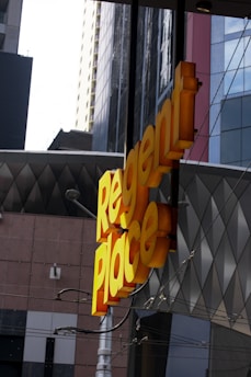 A bright yellow sign with the words 'Regent Place' is prominently displayed against a backdrop of modern urban architecture. The surrounding buildings feature a mix of geometric patterns and glass surfaces.