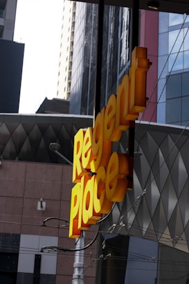 A bright yellow sign with the words 'Regent Place' is prominently displayed against a backdrop of modern urban architecture. The surrounding buildings feature a mix of geometric patterns and glass surfaces.