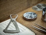 A wooden table holds various tools associated with gem evaluation. A caliper is placed on an open book filled with numbers and gem-related data. A round metal dish contains several cut and polished gemstones. A pair of tweezers is nearby, ready for handling the stones. Metal sieves and a cylindrical container are also present, likely used for sorting and measuring purposes. The overall setting suggests an environment of precision and craftsmanship.