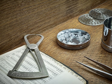 A wooden table holds various tools associated with gem evaluation. A caliper is placed on an open book filled with numbers and gem-related data. A round metal dish contains several cut and polished gemstones. A pair of tweezers is nearby, ready for handling the stones. Metal sieves and a cylindrical container are also present, likely used for sorting and measuring purposes. The overall setting suggests an environment of precision and craftsmanship.
