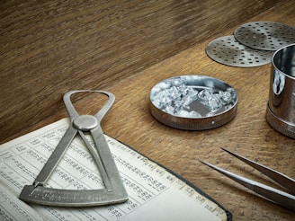 A wooden table holds various tools associated with gem evaluation. A caliper is placed on an open book filled with numbers and gem-related data. A round metal dish contains several cut and polished gemstones. A pair of tweezers is nearby, ready for handling the stones. Metal sieves and a cylindrical container are also present, likely used for sorting and measuring purposes. The overall setting suggests an environment of precision and craftsmanship.
