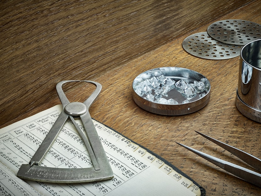 A wooden table holds various tools associated with gem evaluation. A caliper is placed on an open book filled with numbers and gem-related data. A round metal dish contains several cut and polished gemstones. A pair of tweezers is nearby, ready for handling the stones. Metal sieves and a cylindrical container are also present, likely used for sorting and measuring purposes. The overall setting suggests an environment of precision and craftsmanship.