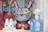 A store display window features several designer toys, including a large, menacing figure with a big grin and sharp teeth, reminiscent of a cartoon character with exaggerated features. Surrounding it are other smaller figures, including a white toy doll and a red and white toy shaped like a rabbit. A blue box labeled 'Mega Munny' contains a blank, customizable figurine.