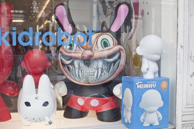 A store display window features several designer toys, including a large, menacing figure with a big grin and sharp teeth, reminiscent of a cartoon character with exaggerated features. Surrounding it are other smaller figures, including a white toy doll and a red and white toy shaped like a rabbit. A blue box labeled 'Mega Munny' contains a blank, customizable figurine.