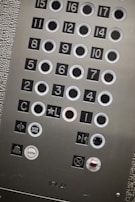 An elevator control panel with multiple round buttons labeled with numbers and symbols. The panel includes floor numbers from 1 to 17, as well as additional buttons for actions like 'Door Open', 'Alarm', and 'Stop'. The surface has a metallic finish with textured and smooth sections. The buttons are arranged in a grid layout, some outlined in white.
