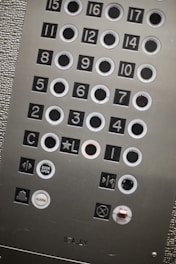 An elevator control panel with multiple round buttons labeled with numbers and symbols. The panel includes floor numbers from 1 to 17, as well as additional buttons for actions like 'Door Open', 'Alarm', and 'Stop'. The surface has a metallic finish with textured and smooth sections. The buttons are arranged in a grid layout, some outlined in white.