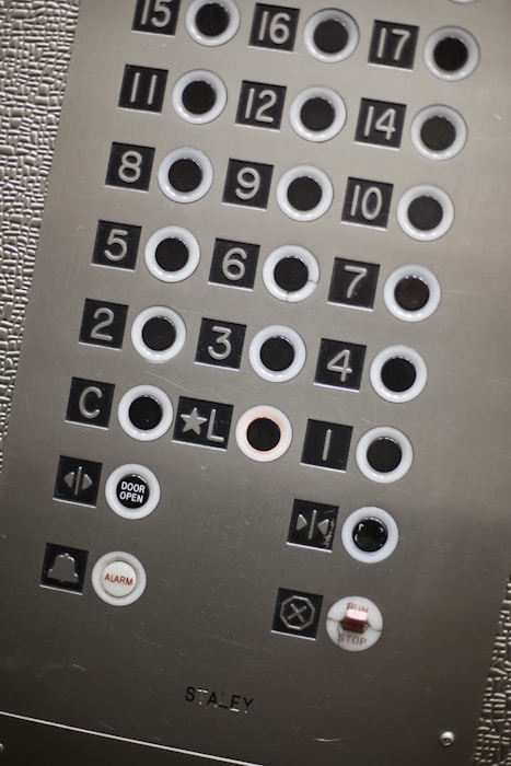 An elevator control panel with multiple round buttons labeled with numbers and symbols. The panel includes floor numbers from 1 to 17, as well as additional buttons for actions like 'Door Open', 'Alarm', and 'Stop'. The surface has a metallic finish with textured and smooth sections. The buttons are arranged in a grid layout, some outlined in white.