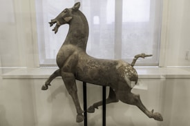 A metal horse sculpture is displayed on a stand in front of a window with translucent blinds. The horse appears dynamic, poised in a mid-stride with its mouth open and ears perked. The surface of the sculpture has a weathered texture, showing patches of discoloration.