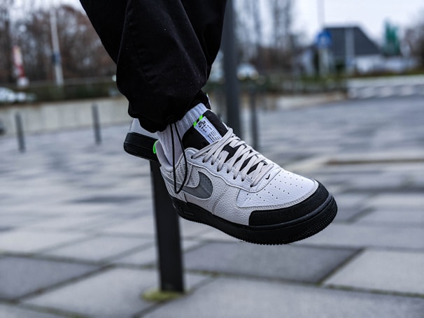 A white and black sneaker with grey accents is shown in mid-air, worn with black pants. The background features an urban setting with a tiled pavement and a blurred view of buildings and trees.