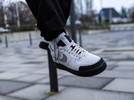 A white and black sneaker with grey accents is shown in mid-air, worn with black pants. The background features an urban setting with a tiled pavement and a blurred view of buildings and trees.