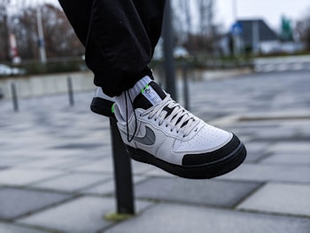 A white and black sneaker with grey accents is shown in mid-air, worn with black pants. The background features an urban setting with a tiled pavement and a blurred view of buildings and trees.