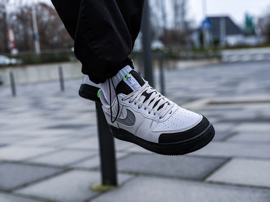 A white and black sneaker with grey accents is shown in mid-air, worn with black pants. The background features an urban setting with a tiled pavement and a blurred view of buildings and trees.