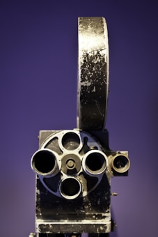 A vintage film projector with a rustic, metal exterior featuring multiple lenses and a large reel holder set against a purple background.