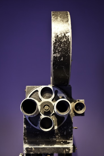 A vintage film projector with a rustic, metal exterior featuring multiple lenses and a large reel holder set against a purple background.