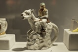 A ceramic figurine depicts a person riding an animal with reptilian features, displayed on a small platform in a showcase. The figure is detailed, with the rider wearing a cap and vest while holding the reins. Surrounding it are other ceramic objects with numbers indicating their placement in a collection.