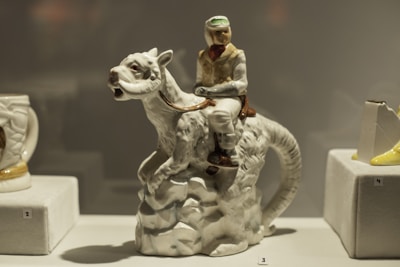 A ceramic figurine depicts a person riding an animal with reptilian features, displayed on a small platform in a showcase. The figure is detailed, with the rider wearing a cap and vest while holding the reins. Surrounding it are other ceramic objects with numbers indicating their placement in a collection.