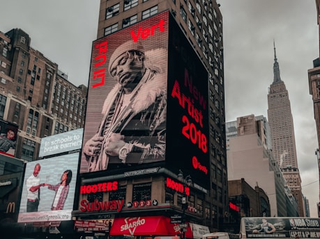 A city street scene with tall buildings featuring large digital billboards. The main billboard prominently displays a monochrome image of a person in fur-trimmed clothing with text highlighting 'New Artist 2018' and 'Li Uzi Vert'. Adjacent billboards include advertisements for schools and fast-food chains, as well as an NBA store. The Empire State Building is visible in the background against a cloudy sky.