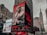 A city street scene with tall buildings featuring large digital billboards. The main billboard prominently displays a monochrome image of a person in fur-trimmed clothing with text highlighting 'New Artist 2018' and 'Li Uzi Vert'. Adjacent billboards include advertisements for schools and fast-food chains, as well as an NBA store. The Empire State Building is visible in the background against a cloudy sky.