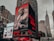 A city street scene with tall buildings featuring large digital billboards. The main billboard prominently displays a monochrome image of a person in fur-trimmed clothing with text highlighting 'New Artist 2018' and 'Li Uzi Vert'. Adjacent billboards include advertisements for schools and fast-food chains, as well as an NBA store. The Empire State Building is visible in the background against a cloudy sky.