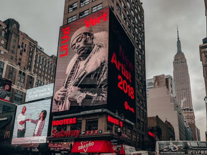 A city street scene with tall buildings featuring large digital billboards. The main billboard prominently displays a monochrome image of a person in fur-trimmed clothing with text highlighting 'New Artist 2018' and 'Li Uzi Vert'. Adjacent billboards include advertisements for schools and fast-food chains, as well as an NBA store. The Empire State Building is visible in the background against a cloudy sky.
